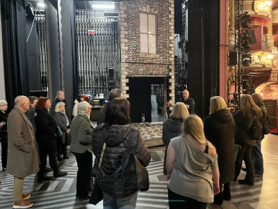 A group of Friends Members stood on stage on the set of Hairspray listening to the company manager give a talk about the show.